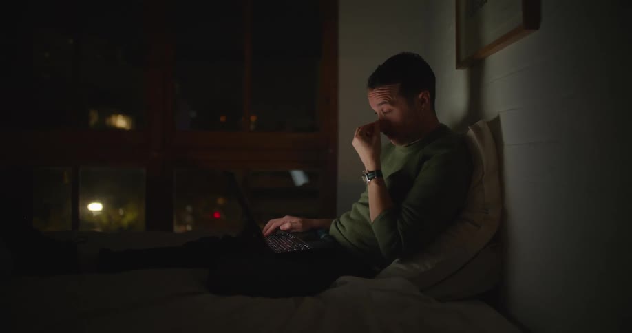 Man with mustache and green sweatshirt sits on bed and uses laptop in dark room