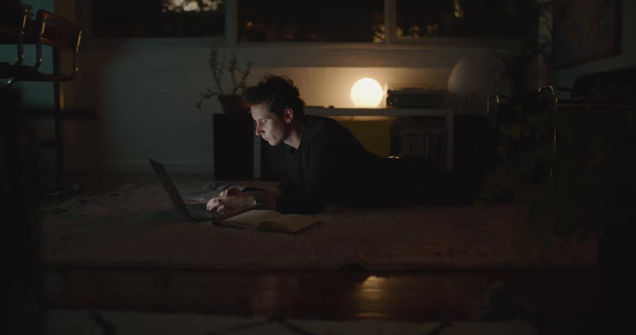 A man is looking at some paperwork then back to typing on his laptop as he sits in a darkened living room