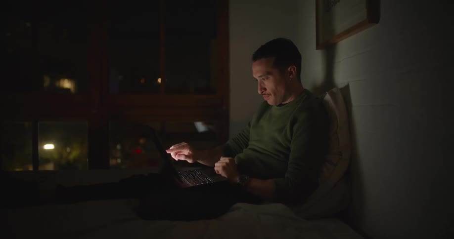 A man is lying on a bed in a dark room typing on a laptop computer