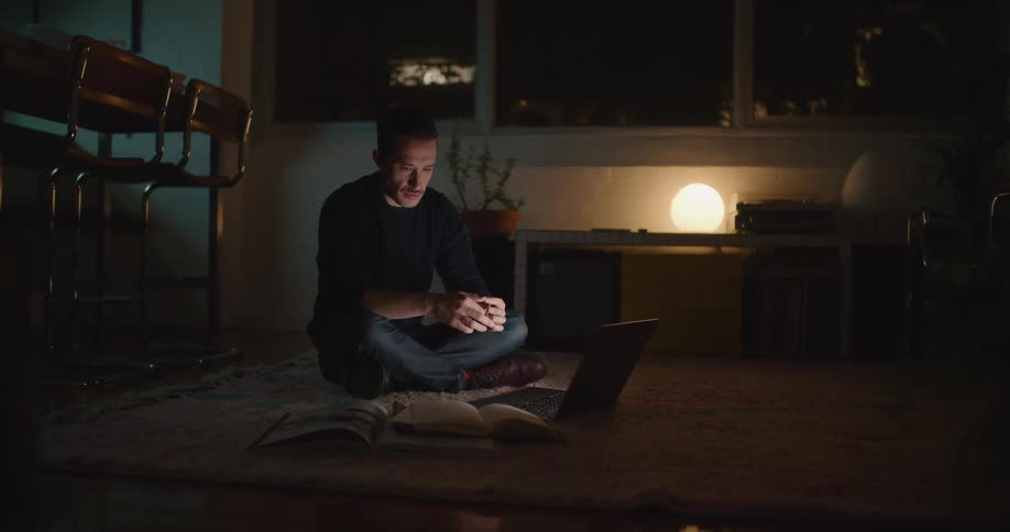 A man sitting cross legged on his living room floor with his laptop and books