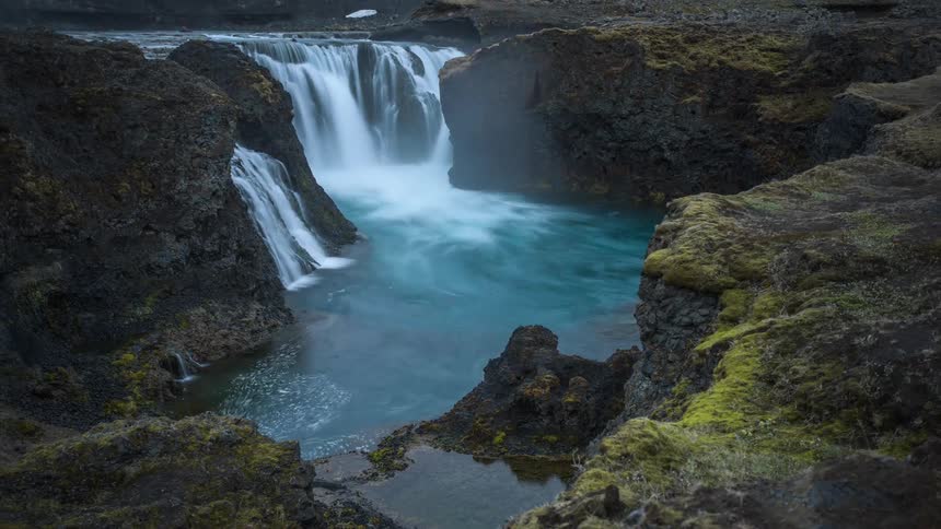 Waterfalls are flowing into a stream among moss covered rocks and there is snow on the ground near the water