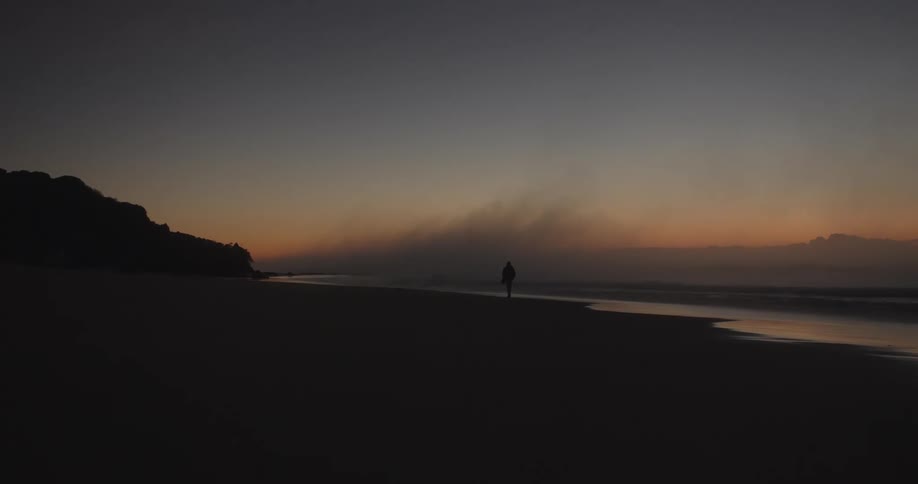 A person walks along an empty beach at sunset