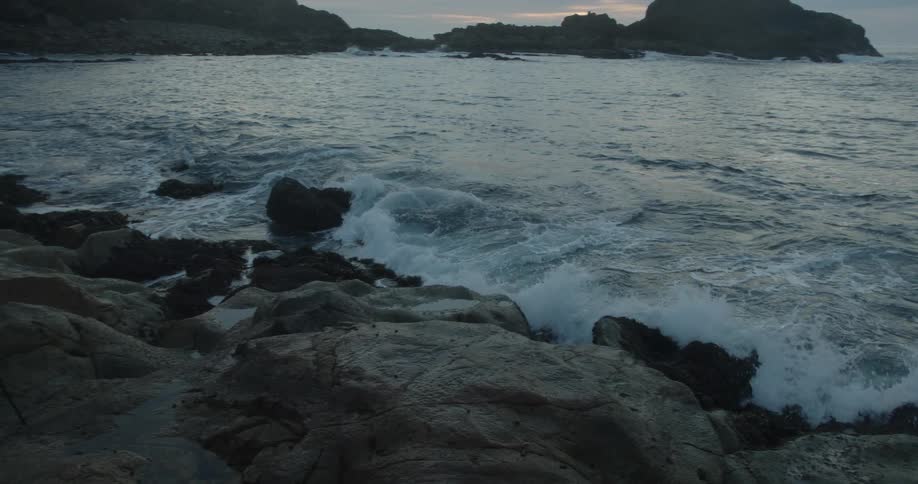 A man sits on a rocky beach with a camera on a tripod as waves roll in