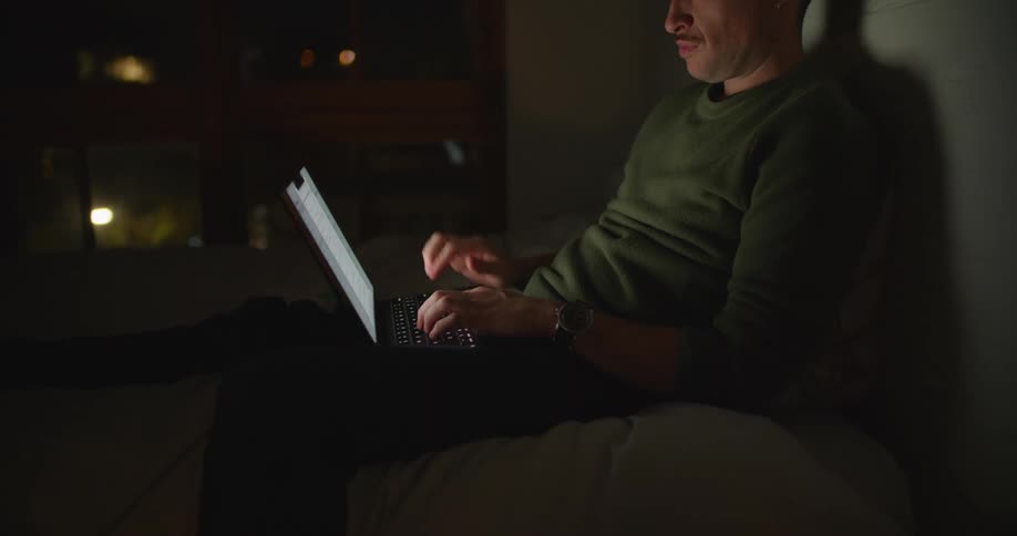 A man sitting up leaning against a wall working on his laptop at night
