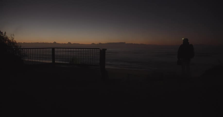 A man walks toward the ocean wearing a head lamp his breath visible in the light