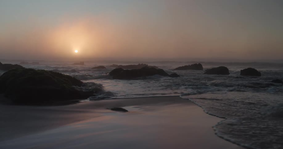 Waves splash against rocks as they roll into shore and over sand on the beach as the sun sets in a hazy sky in the distance