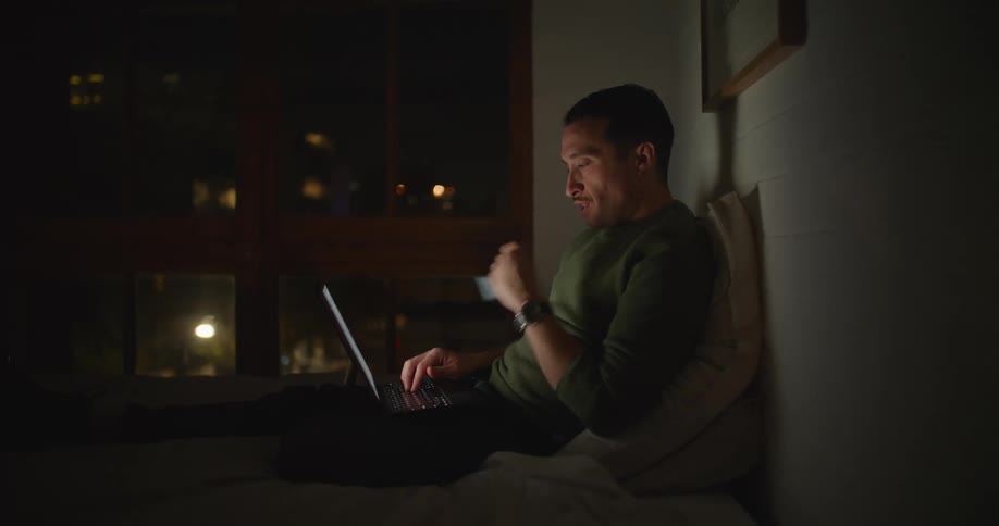 A man is sitting in the dark against a wall typing on his computer