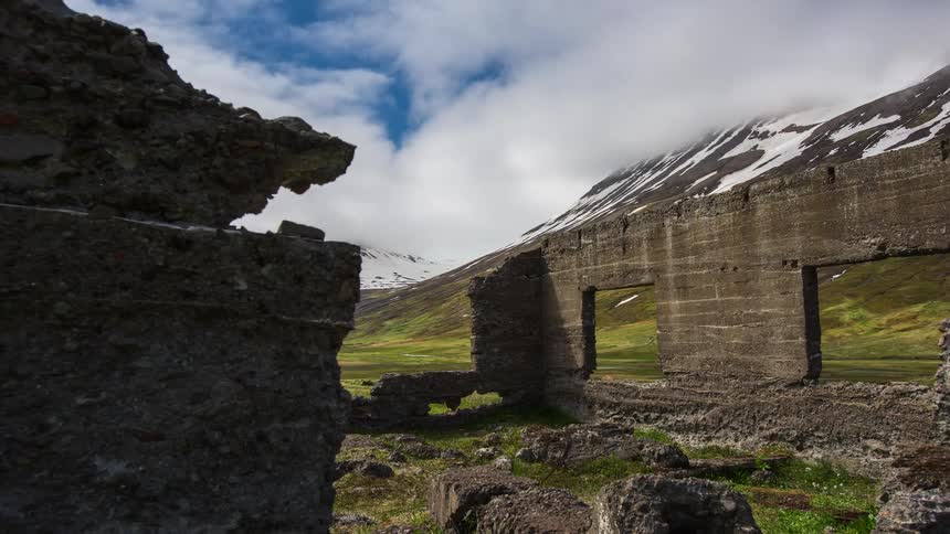 Ruins of a wall sit near mountains with snow on them as clouds move across the sky