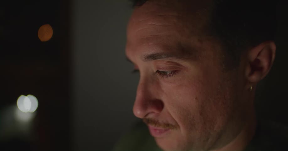 A man wipes his face as he sits in the dark with a light reflecting off of him