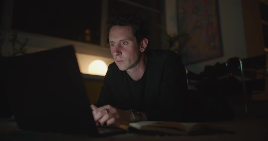 A man references a book while typing on a laptop as he lies on the floor indoors at night