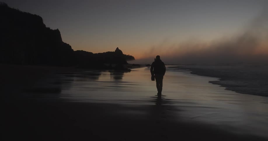 The silhouette of a person walking along a beach while carrying a flashlight at sunset