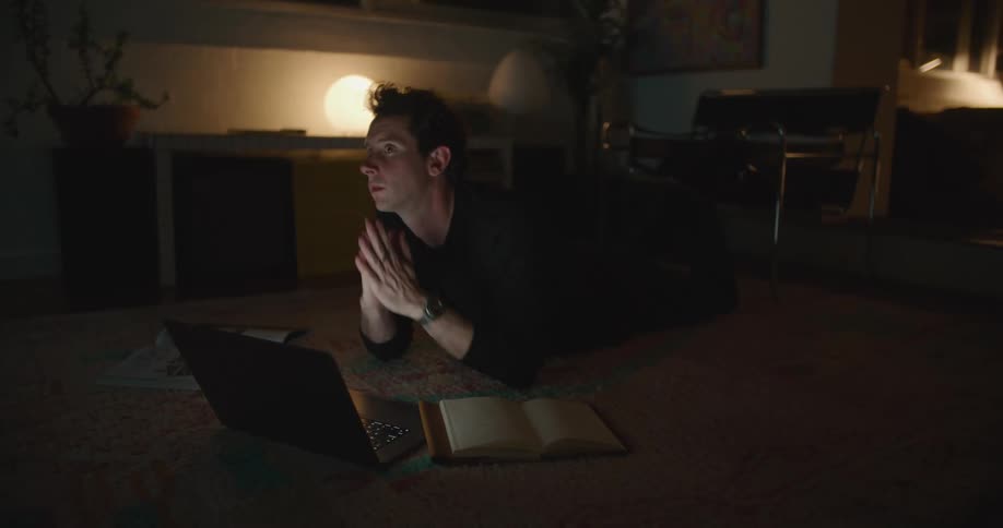 A man is lying on the floor of a dimly lit room and typing on a laptop computer as he looks at a book and then stops to think