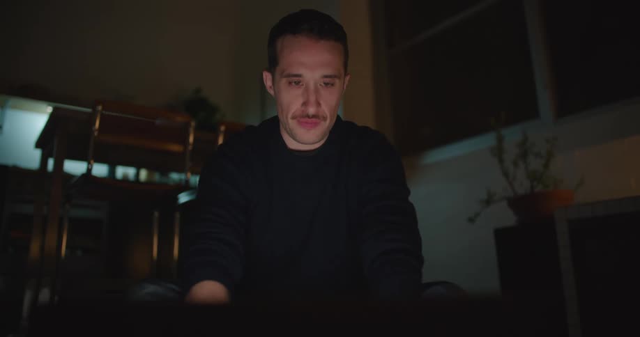 Man sits in dark room thinking and typing on computer