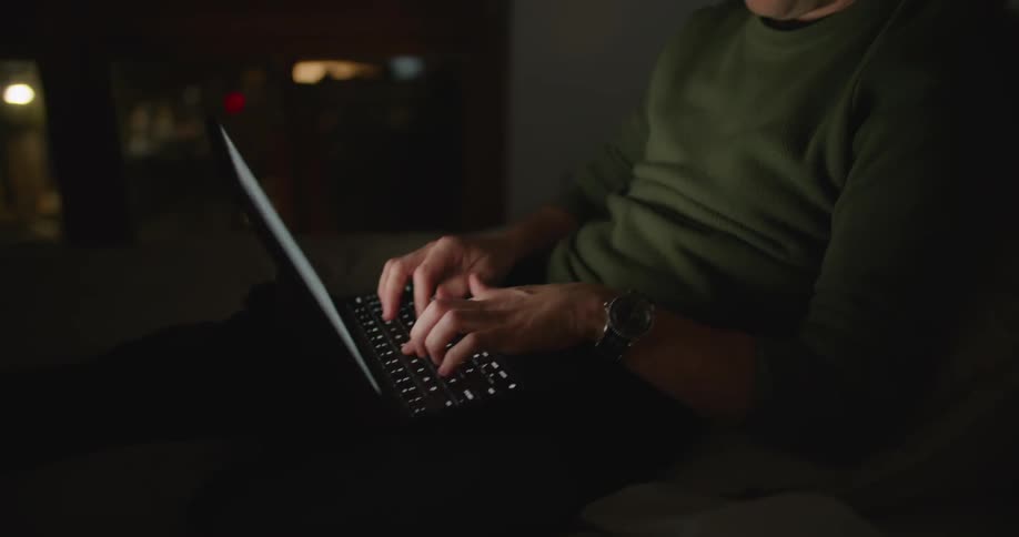 Man with mustache and green sweatshirt types on laptop in dark room