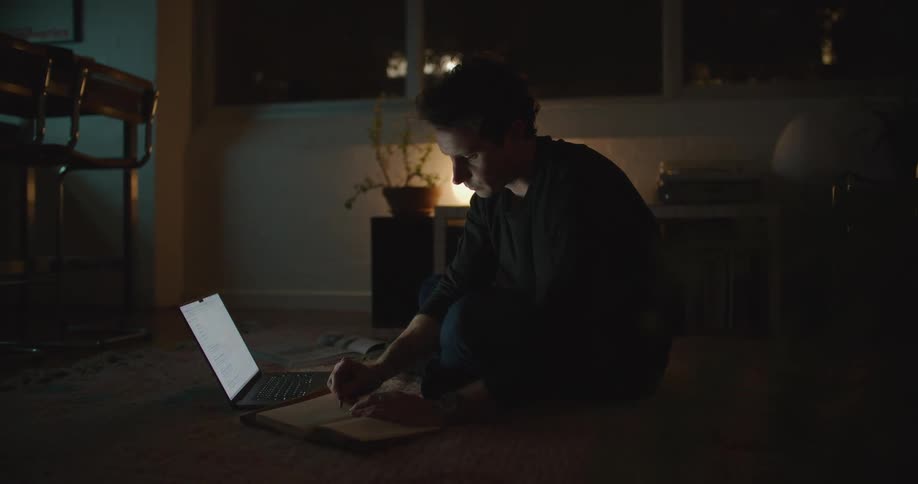A man is sitting on the floor of a dimly lit room and looking at a laptop computer as he writes in a notebook
