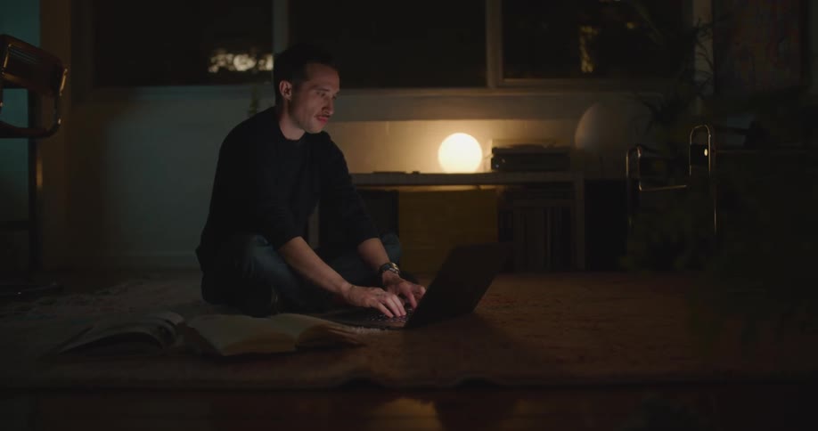 A man is sitting in a room with low light while he types on a laptop and looks to the side