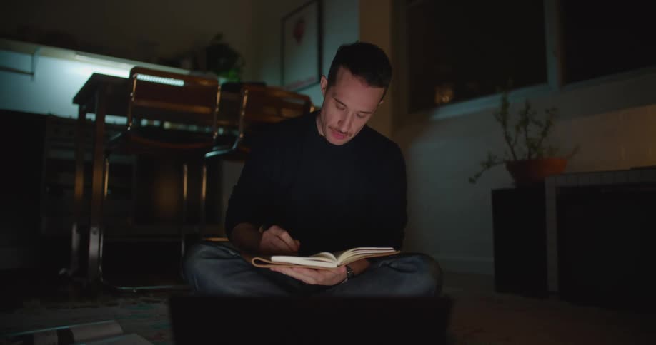 A man consults a book while working on his laptop at home
