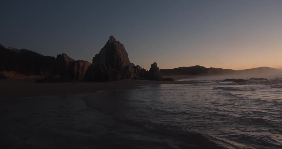 Ocean waves come to shore on a sandy beach with large rocks and a think layer of fog