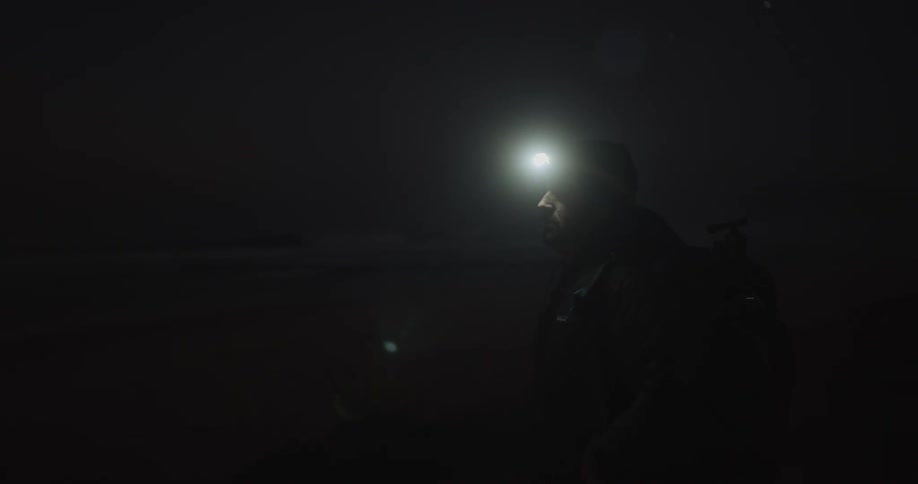 A man wearing a lit headlamp stands then moves slowly in the dark