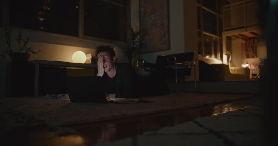 A man is laying on the floor looking at a book and his laptop