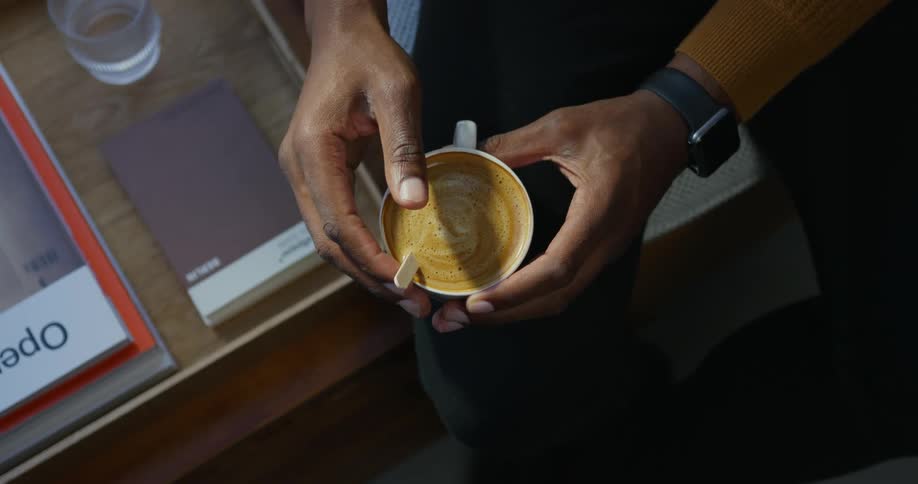A man sitting on a bench next to a tray with books stirs his coffee and lifts it to drink
