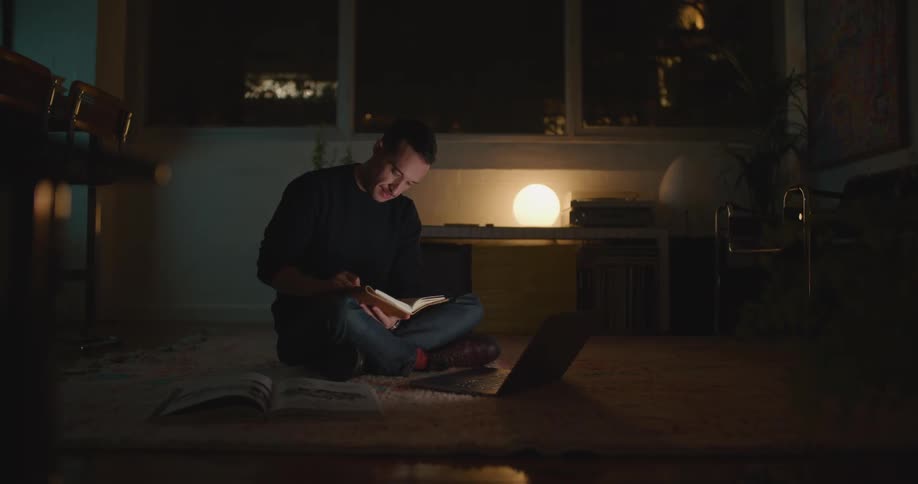 A man sitting on the floor working on a laptop picks up a book for reference