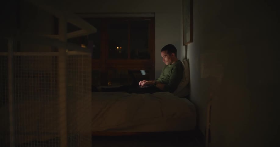 Man sitting against pillows on bed types on laptop in dark room near window showing night