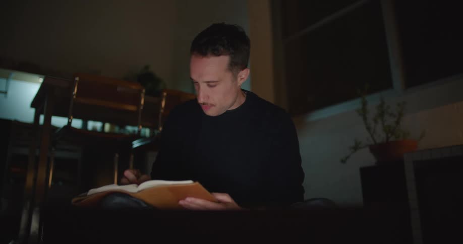 A man is sitting in a dark room writing in a notebook as he looks at a computer