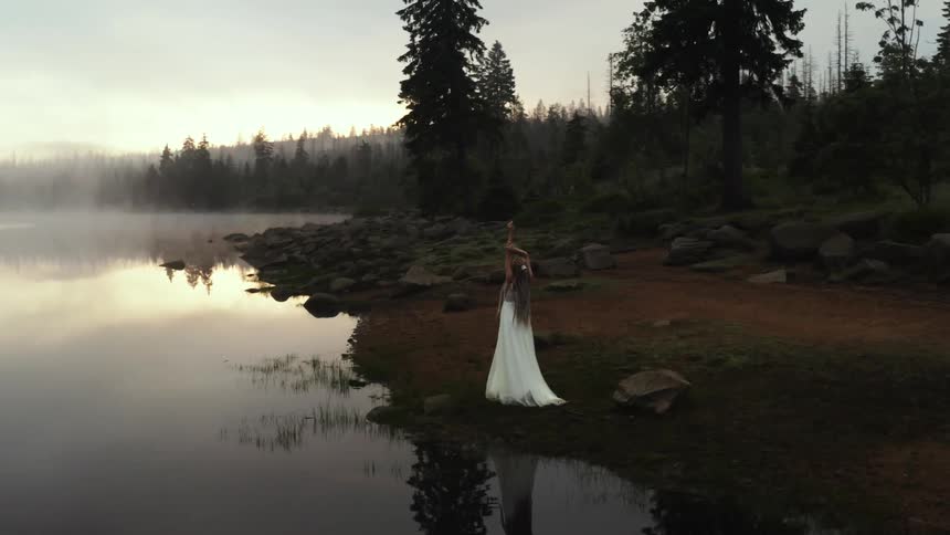 A woman wearing a long white dress is standing at the edge of a lake with her arms stretched upward as fog rises over the water and a forest is in the distance