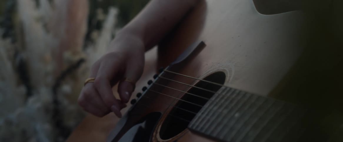 A man wearing gold rings on his finger and thumb is outside strumming an acoustic guitar
