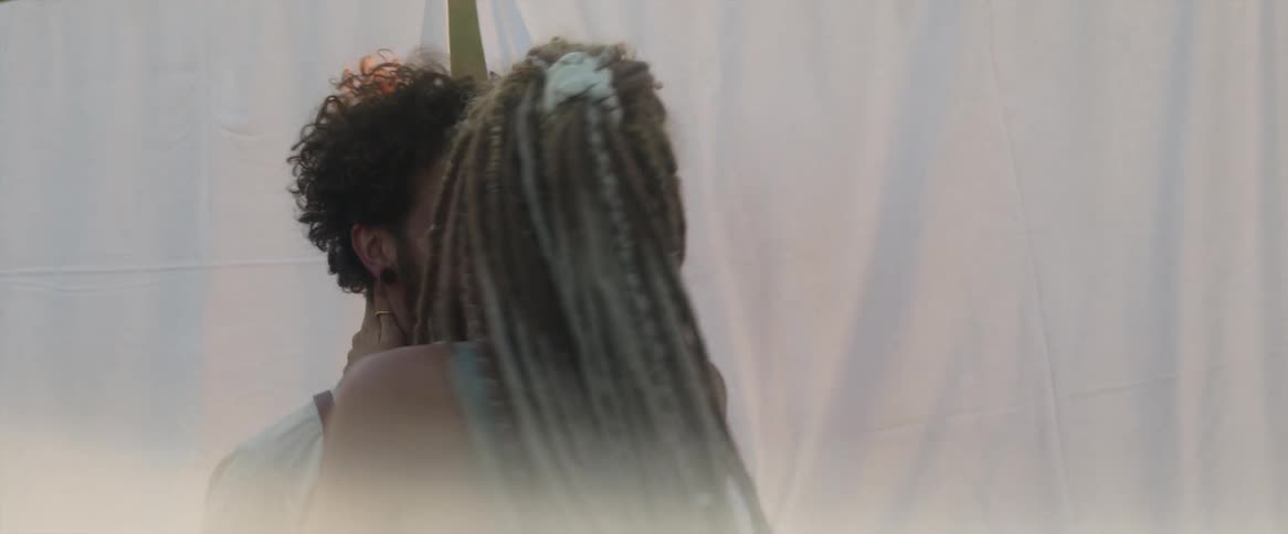A man and a woman are standing in a field among sheets hanging and she puts her hands around his neck and looks as if she will kiss him