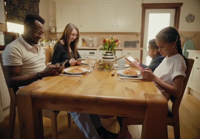 Family sits around table at meal time ignoring food and using their mobile devices