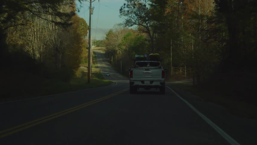 A white truck is traveling on a road in a rural neighborhood passing many trees and a house