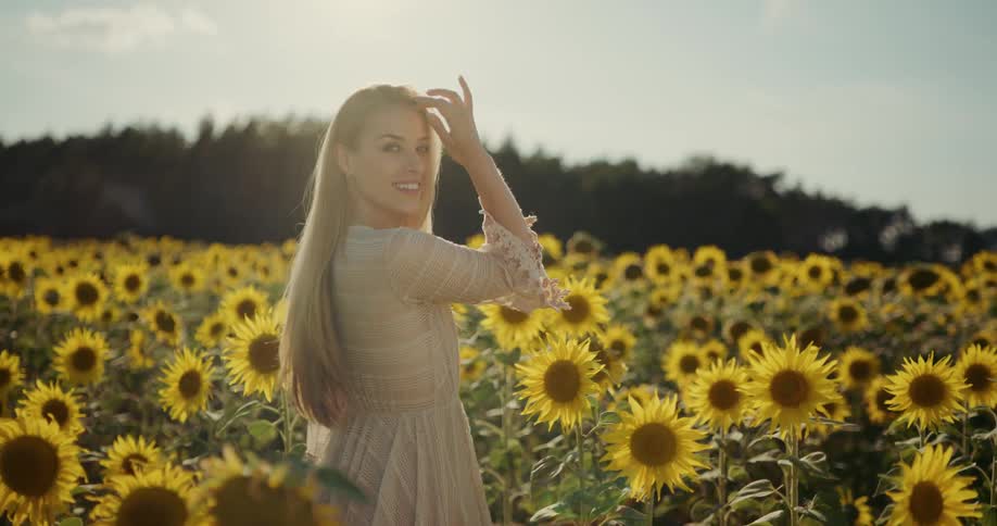 A woman walking through a field of sunflowers turns around to smile into the camera for a moment before continuing onward