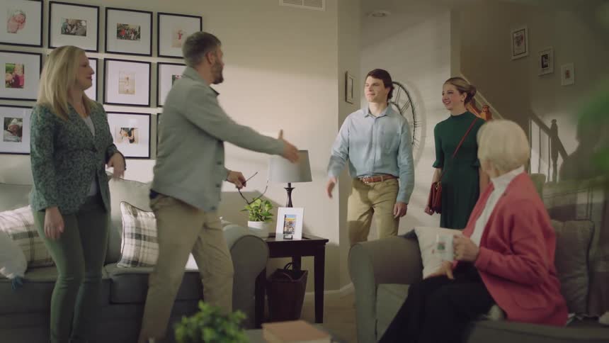 A group of people are in a living room talking when a young couple walks in and greets them