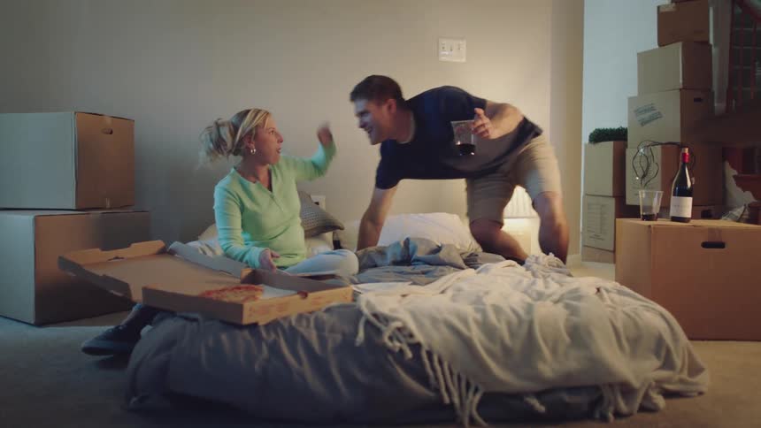 A man and woman are in a house with a lot of moving boxes and they are drinking wine and eating pizza on the mattress