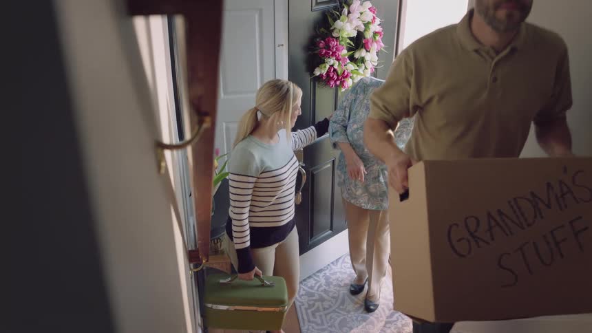 A man is carrying a box up stairs inside a house as a woman helps their grandmother up the stairs
