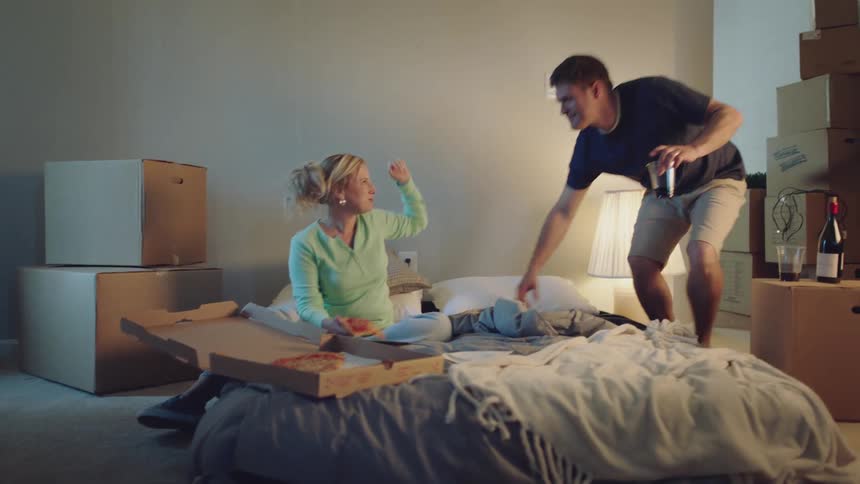 A woman eating pizza raises her arm joyfully in a bedroom full of moving boxes and her husband joins her with wine