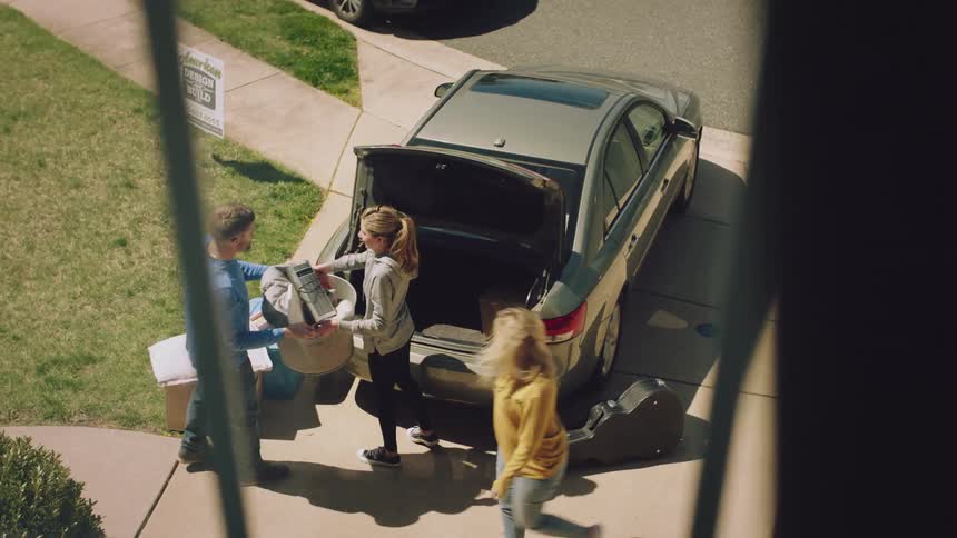 A man carries a basket of electronics and clothing to a pair of women packing things into the trunk of a car parked in a driveway