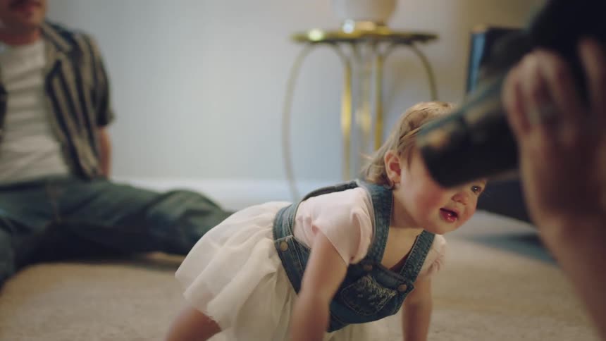 A small child is crawling on the floor away from her father to her mother who is holding a camera