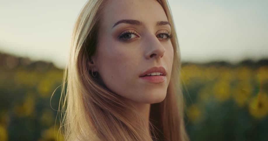 A woman with long blonde hair is standing outside in a field of sunflowers with her eyes closed and she opens them and turns to the camera and smiles