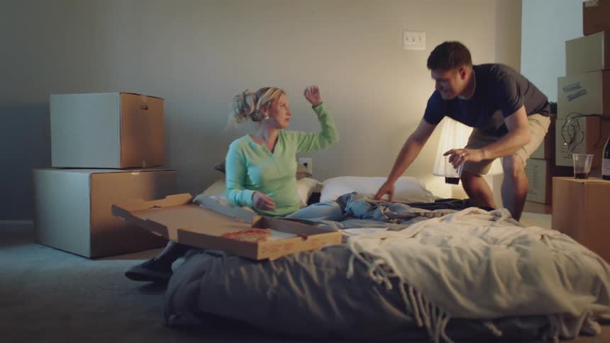 A man and woman are sitting on a mattress on the floor eating pizza and having a good time