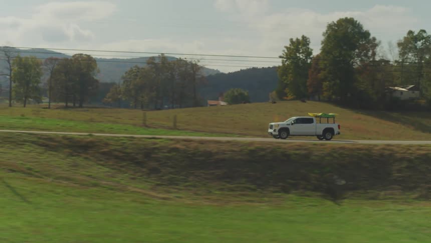 A pickup truck is driving down the road past farmhouses near a forested mountain range