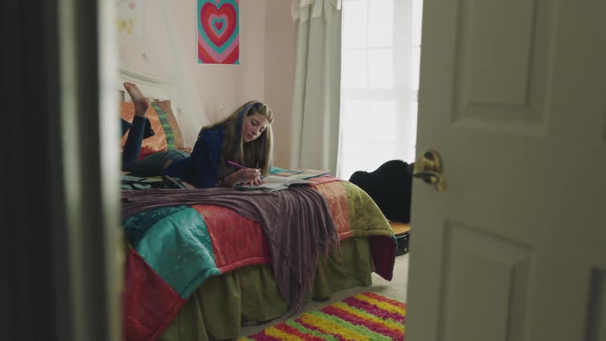 A teenager is laying down on her bed and doing homework