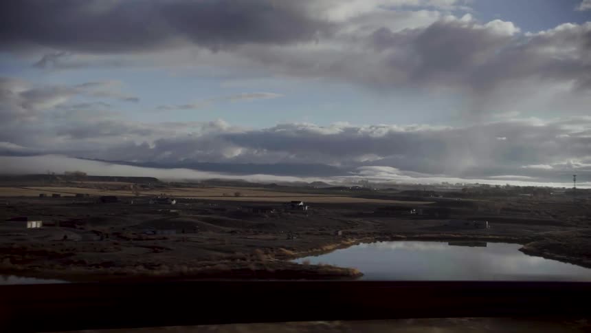 Traveling past open land with farmhouses and a pond with sky and clouds reflected on the water surface