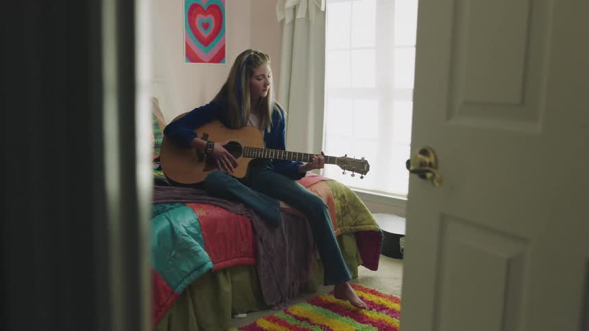 A teenage girl playing guitar while sitting on her bed