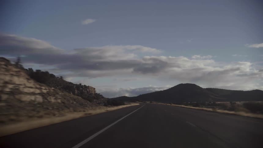 Asphalt topped highway passes low mountains and cliffs on cloudy day
