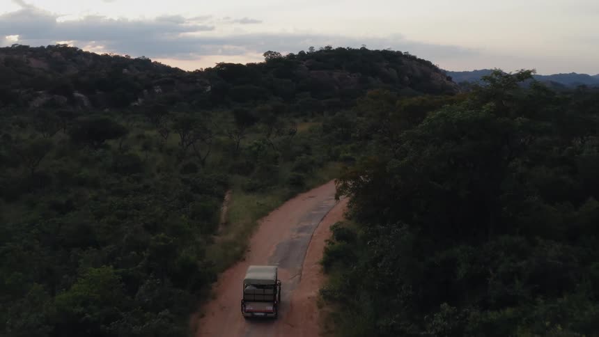 An open truck travels on a narrow dirt and paved road through lush trees and hills