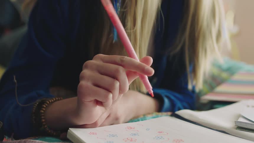 A teenage girl is lying on her bed and writing in a notebook with a pink marker