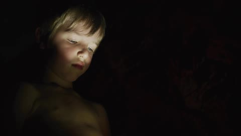 A little boy is standing in the darkness as the light from a flashlight shines on his face
